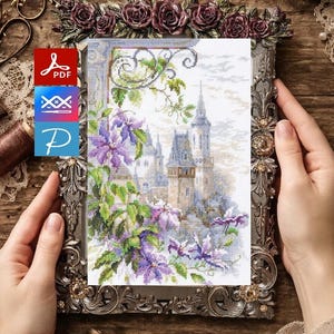 Könnte beinhalten: Ein gerahmtes Kreuzstichbild einer Burgszene mit lila Blumen und grünen Ranken. Der verzierte Rahmen hat ein Blumendesign. Das Bild wird von Händen gehalten. Der Hintergrund ist eine Holzoberfläche mit Nähutensilien.