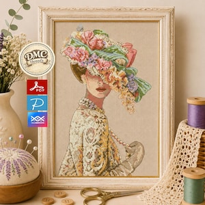 Puede incluir: Retrato de punto de cruz de una mujer con un gran sombrero floral y un collar de perlas, enmarcado en un marco de madera color crema. Suministros de costura rodean la imagen, incluyendo carretes de hilo y un alfiletero.