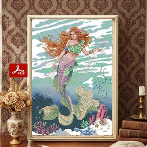 Puede incluir: Una obra de arte en punto de cruz enmarcada representa una sirena con cabello rojo y cola lavanda. Está rodeada de plantas acuáticas y corales en tonos verdes, azules y rosas. La obra está en un marco dorado y sobre un fondo estampado.