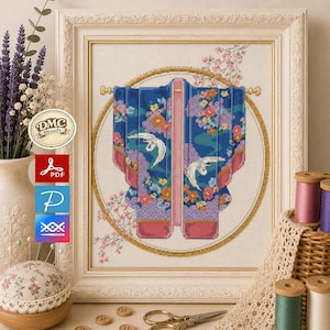 Puede incluir: Patrón de punto de cruz de un kimono azul con diseños florales y de grullas, enmarcado en un marco blanco adornado. Suministros de artesanía, incluyendo carretes de hilo, botones y un alfiletero, rodean la obra de arte. El logotipo de DMC threads es visible.