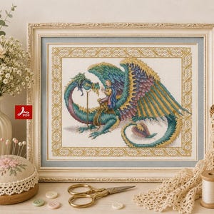 Op de afbeelding: Een ingelijst kruissteek borduurwerk met een grote blauwgroene en gouden draak met een ruiter op de rug, omringd door een geometrische gouden rand. Het kunstwerk is ingelijst in een lichtgekleurde, sierlijke lijst met een blauwe passe-partout.
