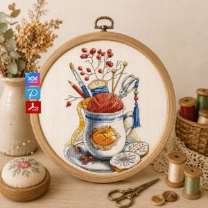 Könnte beinhalten: Eine Kreuzstich-Stickerei in einem Holzrahmen zeigt eine Tasse gefüllt mit Garn, Nadeln und Schere. Ein Fuchs befindet sich auf der Tasse. Beeren und Kekse sind ebenfalls im Design enthalten. Ein Nadelkissen, Garnrollen und eine Schere befinden sich im Vordergrund.