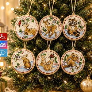 Könnte beinhalten: Sechs kreisförmige Kreuzstich-Ornamente mit Waldtieren wie einem Reh, Waschbären, Hasen und Streifenhörnchen in verschneiten Landschaften. Jedes Ornament ist mit einer goldfarbenen Kordel umrandet und hängt an einer passenden goldenen Schnur.