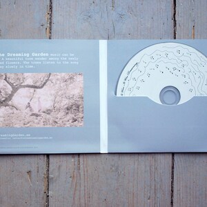 May include: A CD case with a light blue background. The front cover features a black and white photo of a forest with two people walking through it. The text "In the Dreaming Garden" is printed in black at the top of the cover. The back cover features the text "www.dreaminggarden.se".