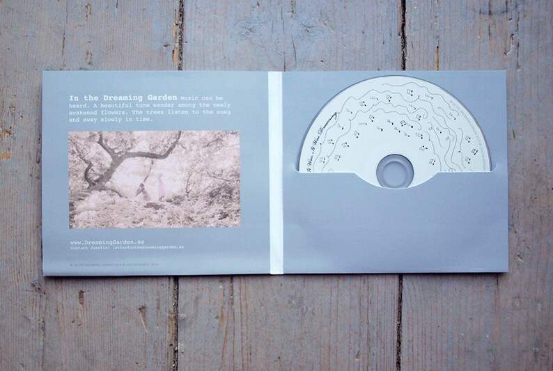 May include: A CD case with a light blue background. The front cover features a black and white photo of a forest with two people walking through it. The text "In the Dreaming Garden" is printed in black at the top of the cover. The back cover features the text "www.dreaminggarden.se".