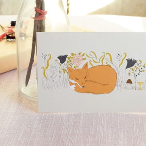 Puede incluir: Una tarjeta de felicitaci&oacute;n con una ilustraci&oacute;n dibujada a mano de un zorro durmiendo en un campo de flores. El zorro es naranja y se acurruca con la cola envuelta alrededor de su cuerpo. Las flores son de varios colores, incluyendo rosa, amarillo y azul.