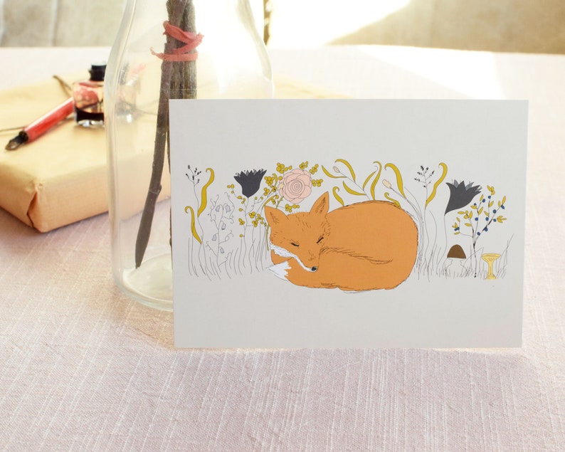 Puede incluir: Una tarjeta de felicitaci&oacute;n con una ilustraci&oacute;n dibujada a mano de un zorro durmiendo en un campo de flores. El zorro es naranja y se acurruca con la cola envuelta alrededor de su cuerpo. Las flores son de varios colores, incluyendo rosa, amarillo y azul.