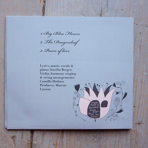 May include: A light blue CD cover with the title "Big Blue Flower" and the track list: "Big Blue Flower", "The Dragonleaf", and "Poem of Love". The cover features a black and white illustration of a flower with pink petals.