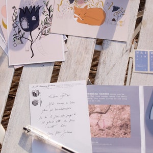Puede incluir: Una postal con una ilustraci&oacute;n dibujada a mano de una casa en una flor con una escalera que lleva a la puerta. La postal est&aacute; sobre una mesa de madera con un bol&iacute;grafo y una caja de CD con una foto de un bosque.