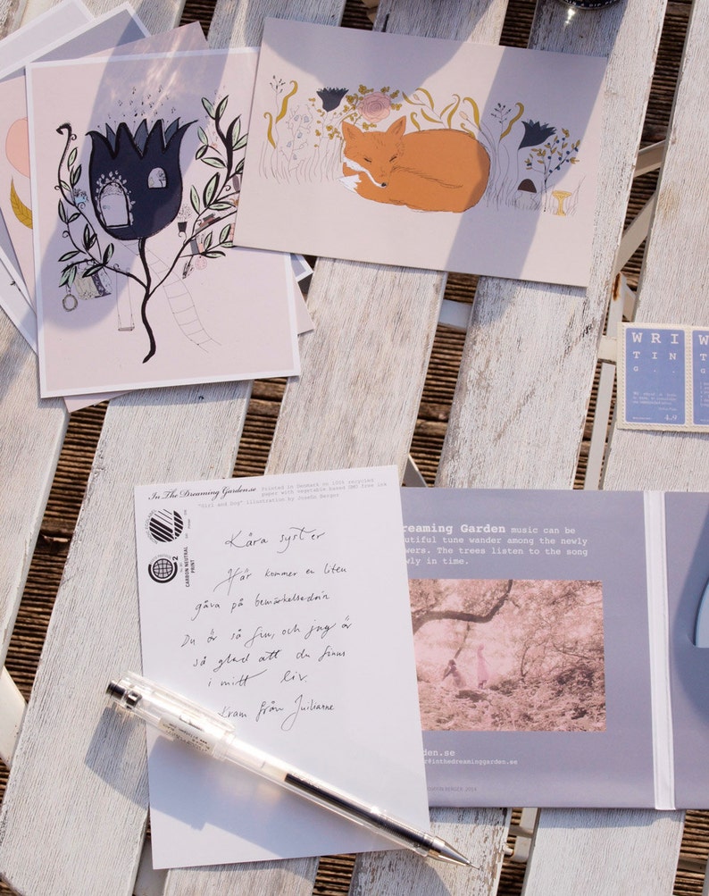 Puede incluir: Una postal con una ilustraci&oacute;n dibujada a mano de una casa en una flor con una escalera que lleva a la puerta. La postal est&aacute; sobre una mesa de madera con un bol&iacute;grafo y una caja de CD con una foto de un bosque.