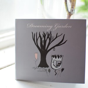 May include: A gray card with the text "Dreaming Garden" printed in white. The card features a black silhouette of a tree with a birdhouse and a tulip with a book inside. The text "Before it was... it was dreaming" is printed below the image.