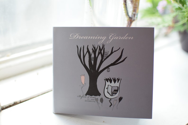 May include: A gray card with the text "Dreaming Garden" printed in white. The card features a black silhouette of a tree with a birdhouse and a tulip with a book inside. The text "Before it was... it was dreaming" is printed below the image.