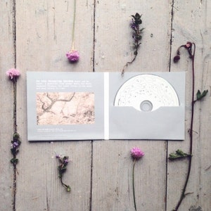 May include: A CD case with a white CD inside. The case is gray and has a white label with the text "In the Breathing Garden, music can be heard. A beautiful tree, under which many of the early enchanting flowers, the forest floor filled in the song and easy beauty in time." The case is surrounded by small pink flowers.