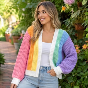 Könnte beinhalten: Eine grob gestrickte Strickjacke mit vertikalen Streifen in Pink, Orange, Gelb, Grün, Blau und Lila. Das vorne offene Kleidungsstück hat weiße Rippbündchen am Ausschnitt, an den Ärmeln und am Saum und wird über einem weißen Oberteil getragen.