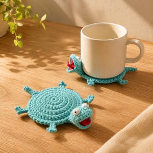 May include: Two light blue crocheted coasters shaped like turtles with round bodies, four small legs, and a head with white eyes and a red mouth. One coaster lies flat, while the other supports a plain, off-white ceramic mug on its back.