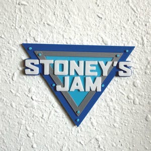 May include: A blue, gray, and white triangular sign with the text "STONEY'S JAM". The sign has a blue border with gray and light blue interior sections. The text is white and in a bold, sans-serif font.
