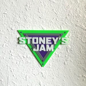 May include: A green, purple, and gray triangular sign with the text "STONEY'S JAM" in white. The sign has a textured surface and is mounted on a white wall. The design features a layered effect with a geometric shape.