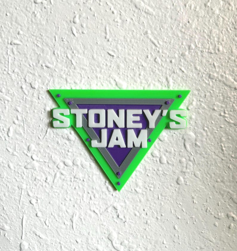 May include: A green, purple, and gray triangular sign with the text "STONEY'S JAM" in white. The sign has a textured surface and is mounted on a white wall. The design features a layered effect with a geometric shape.