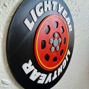 May include: A round, black wall decor piece with the text "LIGHTYEAR" in white, raised lettering. The center features a red wheel design with yellow and gray accents. The design is reminiscent of a tire.