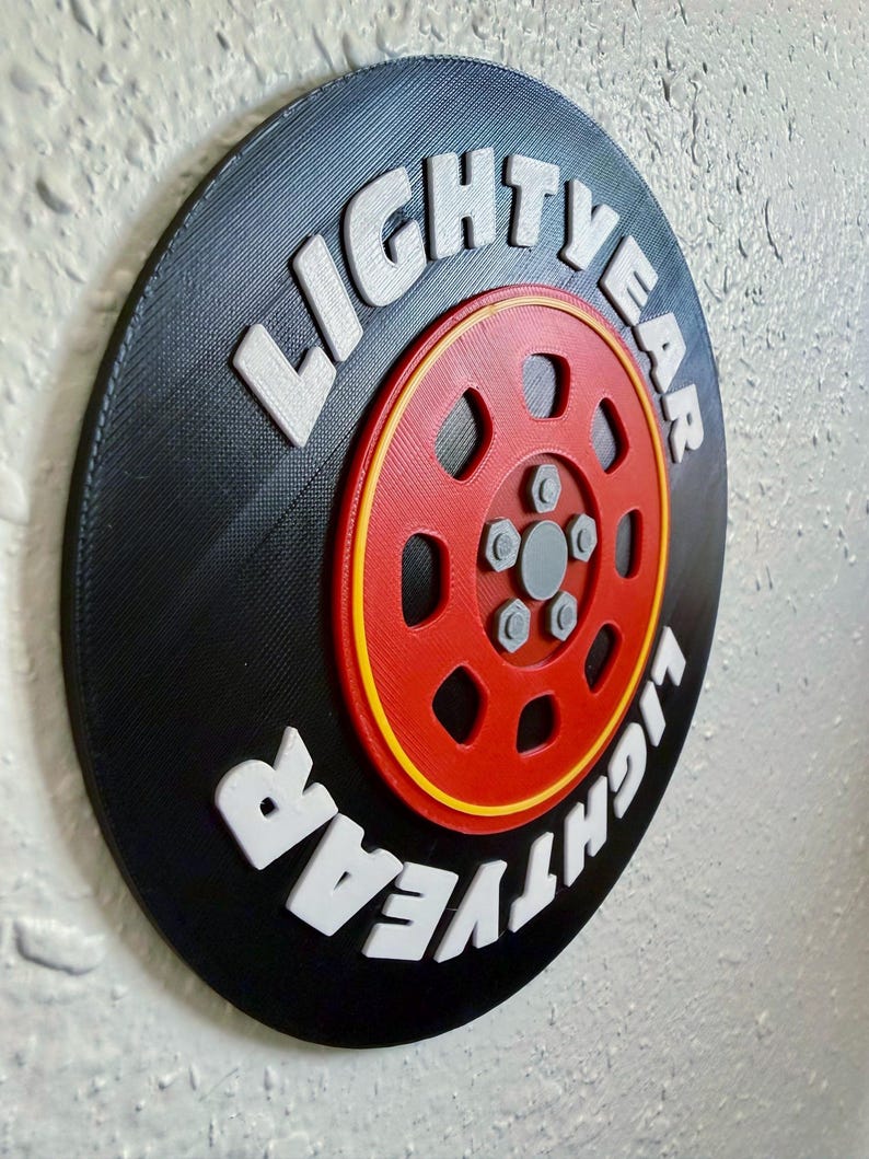 May include: A round, black wall decor piece with the text "LIGHTYEAR" in white, raised lettering. The center features a red wheel design with yellow and gray accents. The design is reminiscent of a tire.