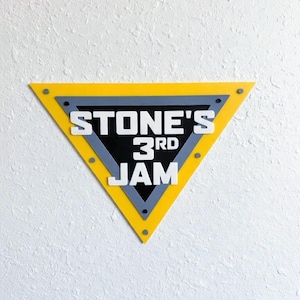 May include: A yellow, gray, and black triangular sign with the text "STONE'S 3RD JAM" in white lettering. The sign is mounted on a white textured wall. The sign has a yellow border, a gray inner border, and a black center.
