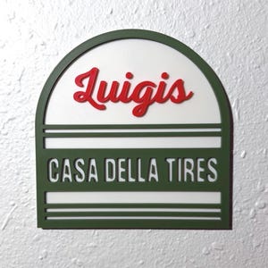 May include: A green and white sign with the words "Luigis" in red script above "CASA DELLA TIRES" in white block letters. The sign has a semi-circular top and is mounted on a textured white wall.