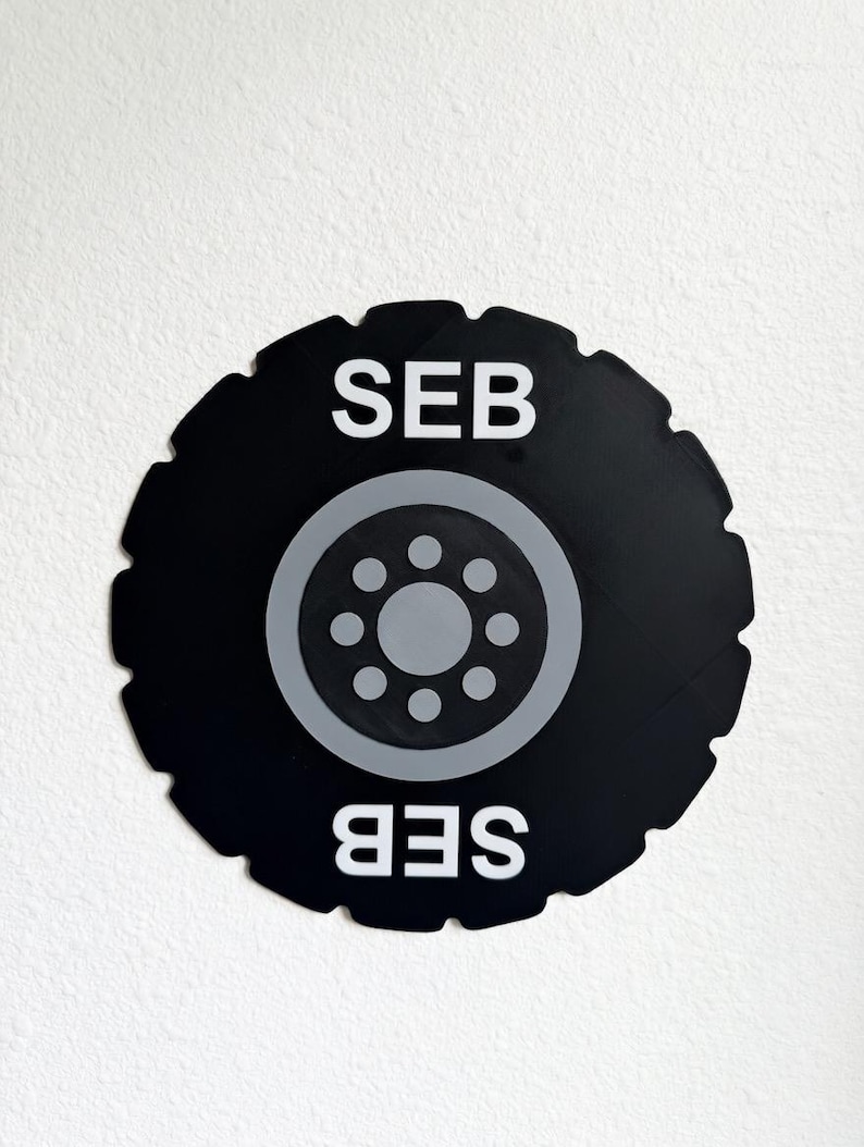 May include: Black circular wall art featuring a tire tread design with a gray wheel center. The letters "SEB" are displayed in white at the top and bottom of the design. The artwork is set against a white background.