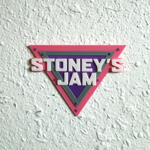 May include: A triangular sign with the text "STONEY'S JAM" in white letters. The sign has a pink border, a gray middle section, and a purple center. Small, round, purple accents are visible on the corners.