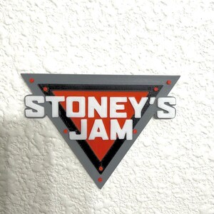 May include: A triangular sign with the text "STONEY'S JAM" in white letters. The sign has a gray border, a red center, and small red accents. The sign is mounted on a textured white wall.