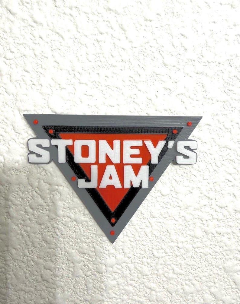 May include: A triangular sign with the text "STONEY'S JAM" in white letters. The sign has a gray border, a red center, and small red accents. The sign is mounted on a textured white wall.