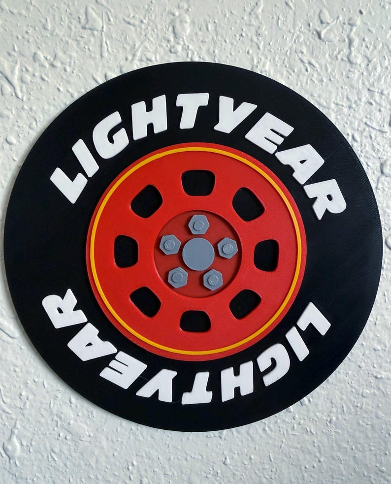 May include: A round black and red decorative piece with the text "LIGHTYEAR" in white. The design features a red wheel with a yellow outline and gray bolts, resembling a tire.