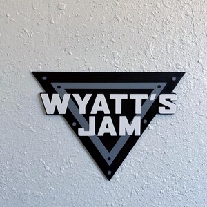 May include: A black, gray, and white wooden sign in a triangular shape. The sign reads "WYATT'S JAM" in bold, white letters. The sign is mounted on a textured white wall.
