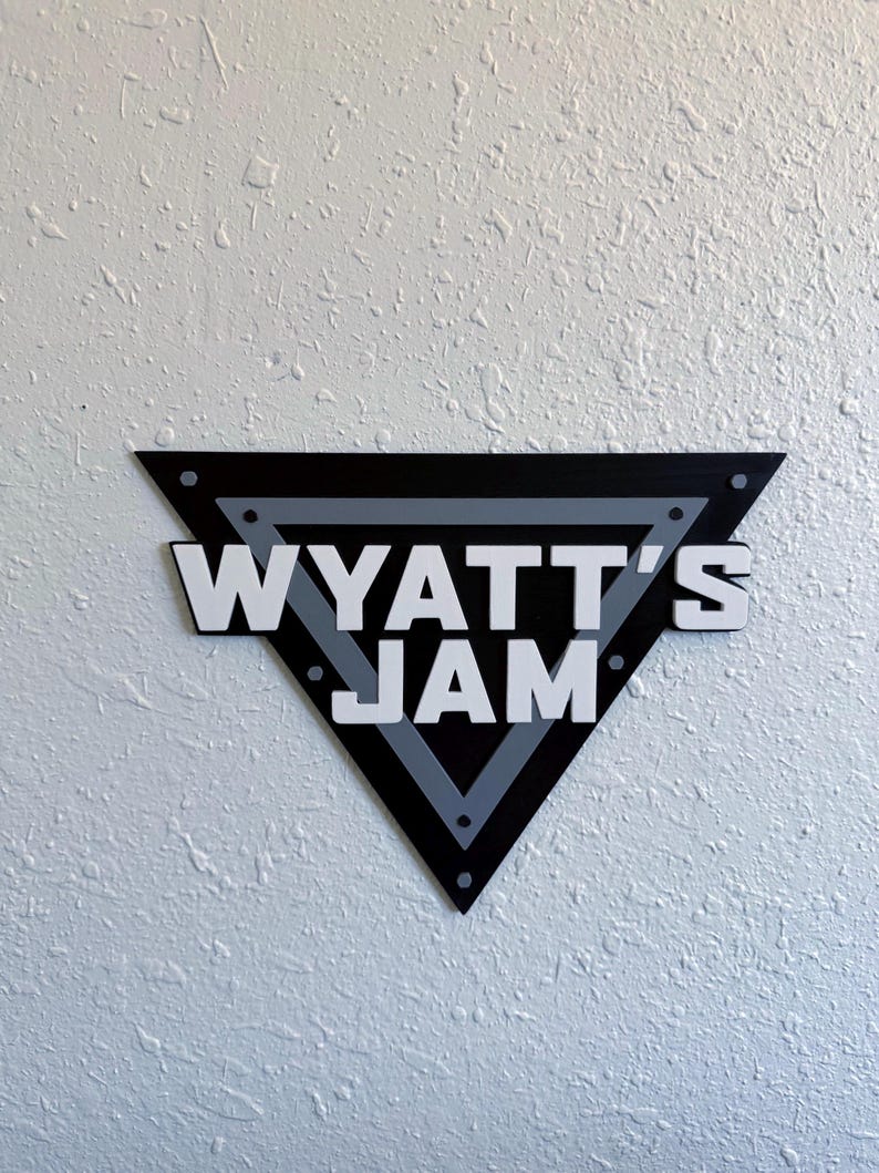 May include: A black, gray, and white wooden sign in a triangular shape. The sign reads "WYATT'S JAM" in bold, white letters. The sign is mounted on a textured white wall.