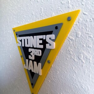May include: A yellow, gray, and black triangular sign with the text "STONE'S 3RD JAM" in white, mounted on a textured white wall. The sign has a layered design with visible screws.