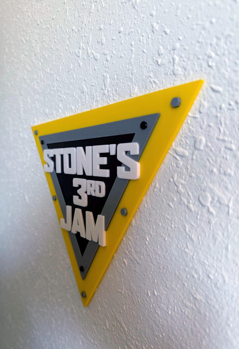 May include: A yellow, gray, and black triangular sign with the text "STONE'S 3RD JAM" in white, mounted on a textured white wall. The sign has a layered design with visible screws.