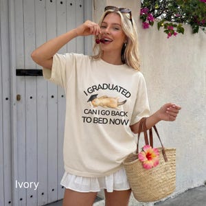 May include: Ivory-colored t-shirt with the text "I GRADUATED CAN I GO BACK TO BED NOW" and a graphic of a cat wearing a graduation cap. The shirt is paired with a white skirt and a straw tote bag with a pink flower.