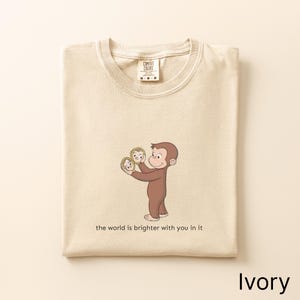 May include: An ivory-colored t-shirt featuring a graphic of a brown monkey holding two small gold-framed mirrors. Below the illustration, the text reads, "the world is brighter with you in it." The shirt has a Comfort Colors brand label at the neck.