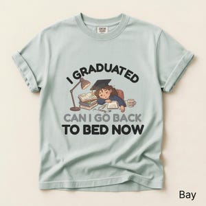 May include: Light blue t-shirt with the text "I GRADUATED CAN I GO BACK TO BED NOW" and a graphic of a student sleeping on books. The shirt has rolled-up sleeves and a crew neck.
