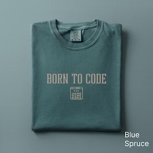 May include: Blue spruce t-shirt with the text "BORN TO CODE" in a light brown color. Below the text is a graphic of a computer screen with code. The shirt is folded and has a crew neck.