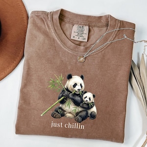 Puede incluir: Una camiseta de manga corta de color marrón con un gráfico de un panda adulto y un bebé panda comiendo tallos de bambú. El texto "just chillin" está impreso en blanco debajo de los pandas. La camiseta tiene una etiqueta Comfort Colors.