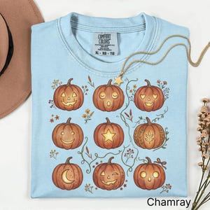May include: A light blue t-shirt featuring a graphic print of nine orange pumpkins with various carved facial expressions and botanical accents. The shirt has a crew neckline and a Comfort Colors brand label printed on the inside of the collar.