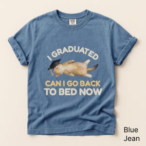 May include: Blue jean-colored t-shirt with rolled sleeves. The shirt features the text "I GRADUATED CAN I GO BACK TO BED NOW" above an illustration of a cat wearing a graduation cap. The cat is lying on its back.