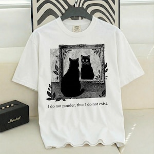 May include: White t-shirt featuring a black cat silhouette looking into a framed mirror, with a reflection of itself. The text "I do not ponder, thus I do not exist" is printed below the image. The shirt has short sleeves.