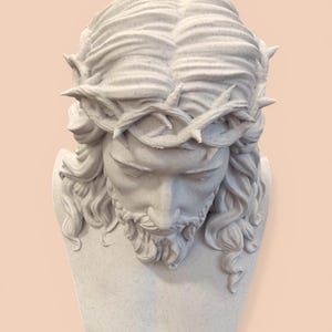 Puede incluir: Escultura de busto blanco que representa una figura con pelo largo, barba y corona de espinas. La escultura tiene superficies texturizadas y está sobre un fondo melocotón claro.