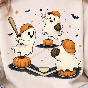 May include: A cream-colored sweatshirt featuring a graphic of three white ghosts wearing orange baseball caps. One ghost holds a wooden bat while standing on a pumpkin, another catches a baseball, and the third crouches on a pumpkin.