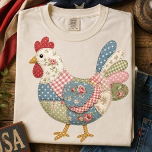 May include: Cream-colored t-shirt featuring a patchwork chicken design. The chicken is composed of various fabric patterns, including floral, polka dot, and gingham. The chicken has a red comb and wattle, and yellow feet. The shirt has a crew neck and short sleeves.