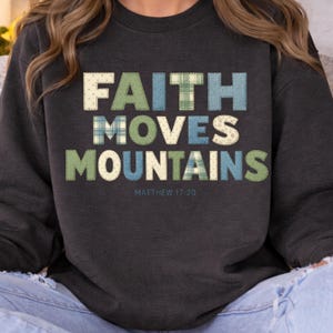 Puede incluir: Sudadera gris oscuro con las palabras "FAITH MOVES MOUNTAINS" en letras de estilo patchwork. El texto es en tonos verdes, azules y crema, con el versículo bíblico Mateo 17:20 debajo. Una prenda superior cómoda e informal.
