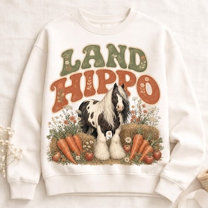 Pode incluir: Uma camisola de gola redonda cor de creme com um gráfico de um cavalo preto e branco num campo de flores silvestres e feno. O texto "LAND HIPPO" está impresso em letras grandes, curvas e de estilo retro sobre o cavalo.
