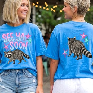 May include: Two women wearing matching blue t-shirts with a raccoon graphic. The front of the shirt says "SEE YA SOON" in pink and green. The back of the shirt features a raccoon and star graphics.