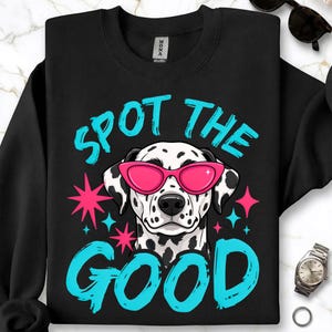 May include: A black crewneck sweatshirt featuring a graphic of a Dalmatian dog wearing bright pink cat-eye sunglasses. The text "SPOT THE GOOD" is printed in a bold, light blue font above and below the dog, accented by pink and blue stars.